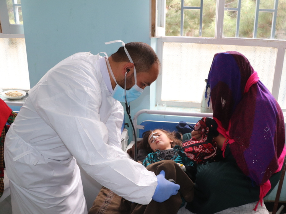 En la foto, un médico de MSF con ropa protectora, se inclina para estar al nivel de la cama de su paciente, una pequeña con sarampión recostada en la coma, cubierta por una manta y con una sonda nasal. Al lado de la pequeña, está su madre.