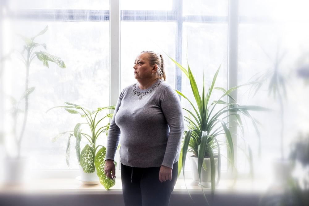 Una mujer de mediana edad, rubia y de tez clara, en medio de una habitación ampliamente iluminada y rodeada de plantas.