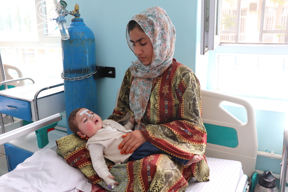 En una cama de hospital, con una ventana y un tanque de oxígeno, está sentada, una madre con hijab tiene a su pequeño en las piernas. El pequeño tiene una sonda en la nariz.