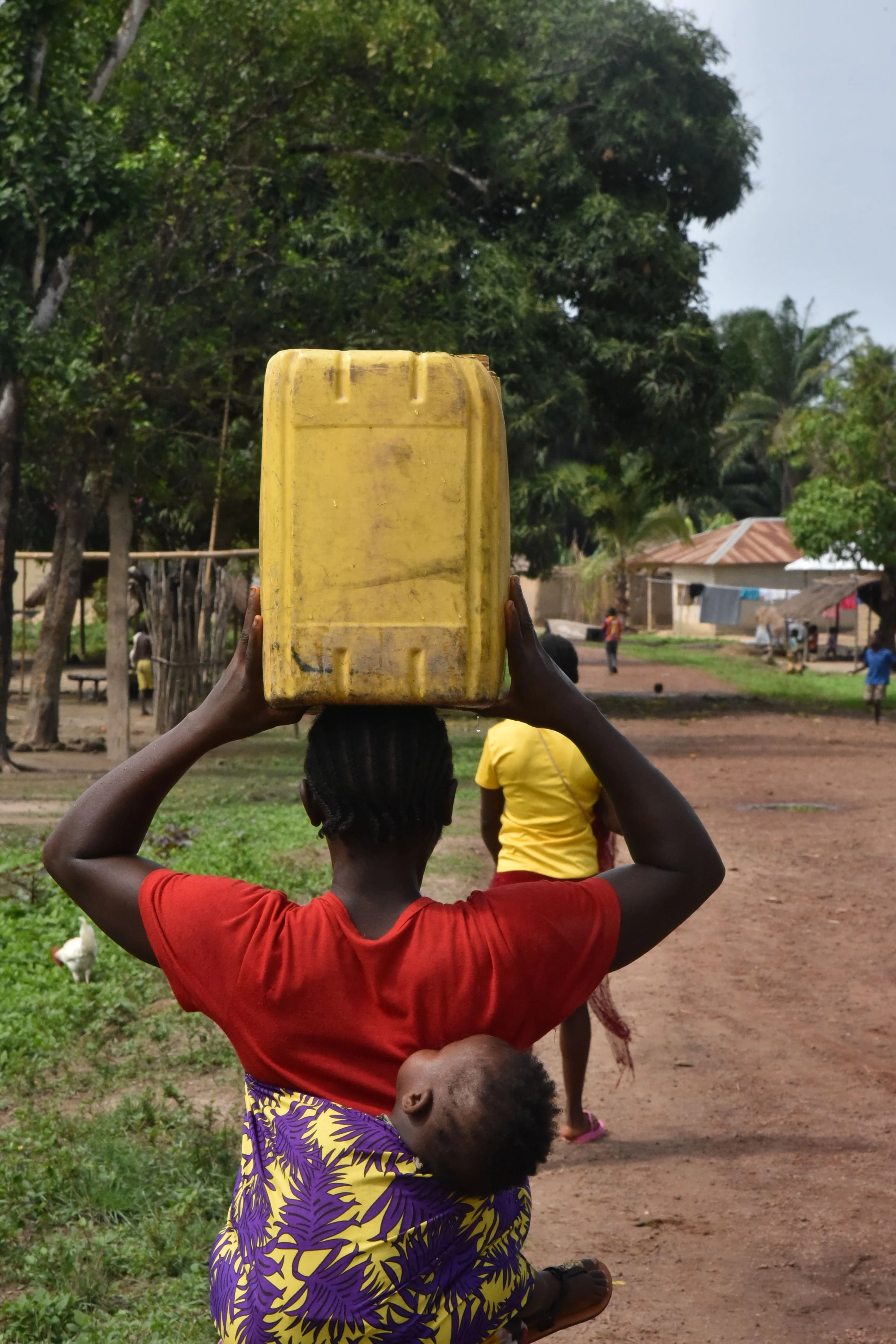 Aminata Bangura lleva un bidón de 20 litros lleno de agua sobre la cabeza y camina hacia su casa con su hijo atado a la espalda en la aldea de Masiperr, distrito de Tonkolili, Sierra Leona. Aminata fue a buscar agua de un pozo perforado por MSF cerca de la Unidad de Salud Periférica (USP) de la aldea de Masiperr, con bomba manual. © Daniel García / MSF