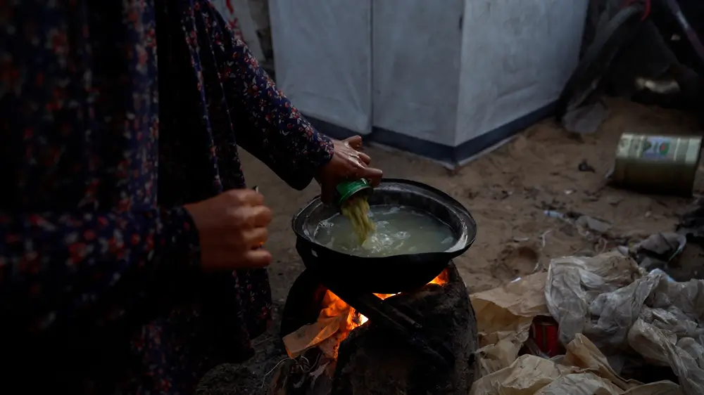 Una madre prepara comida para sus hijos y familia, vertiendo una lata de judías verdes en agua hirviendo sobre un fuego que ella misma enciende. La cantidad de ayuda humanitaria que entra en Gaza alcanza su nivel más bajo en meses.