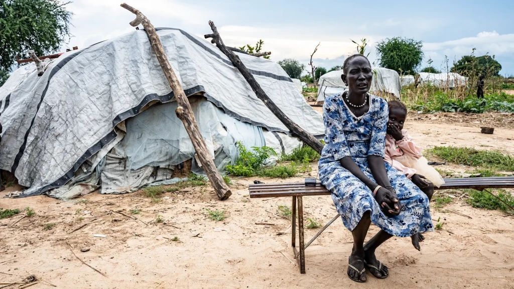 Arual Manyok y su hija Adit Ayuel se sientan en un banco frente a su refugio en un campo de personas desplazadas. Sudán del Sur, octubre de 2024.
