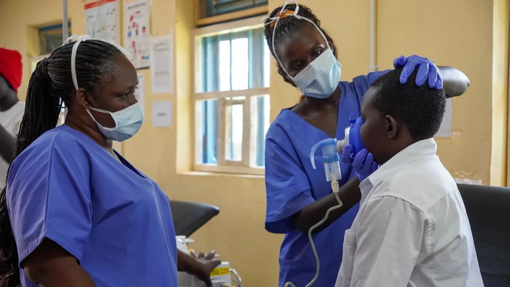 La mentora clínica Florence observa a la enfermera Agnes administrando medicamentos a un paciente a través de un nebulizador en la sala de emergencias.
