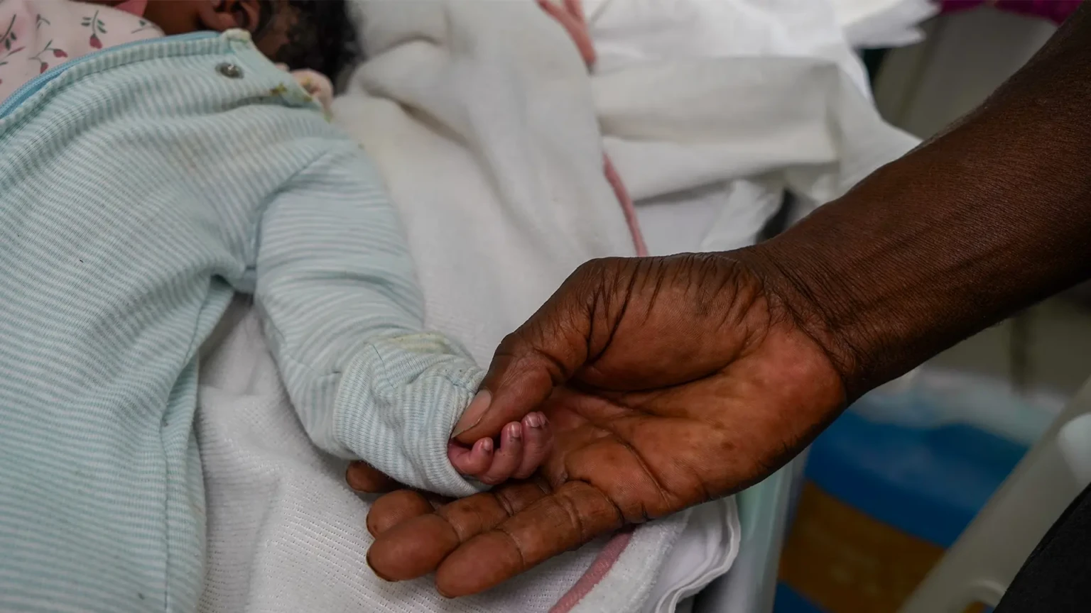 La mano de un recién nacido descansa en la palma de su abuelo, que buscó atención de urgencia para la madre del bebé en la Maternidad Isaïe Jeanty, tras una eclampsia posparto, una complicación que ponía en peligro la vida.