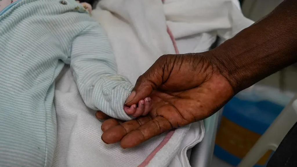 La mano de un recién nacido descansa en la palma de su abuelo, que buscó atención de urgencia para la madre del bebé en la Maternidad Isaïe Jeanty, tras una eclampsia posparto, una complicación que ponía en peligro la vida.