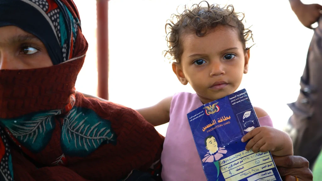 Asmaa Abdo, de 2 años, sostiene su tarjeta de vacunación mientras espera con su madre, Zubaida Ali, durante una actividad de vacunación contra el sarampión en Ateera (2), distrito de Tuban, gobernación de Lahj, Yemen, 5 de mayo de 2026.