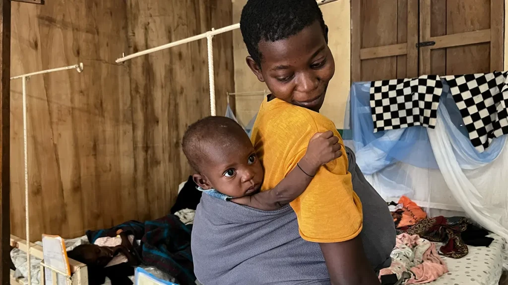 Maombi sostiene a su hija de un año, Wabiwa, a al espalda, sonriéndole frente a la cámara del hospital mientras recibe tratamiento por desnutrición aguda en el hospital general de referencia de Walikale, RDC.